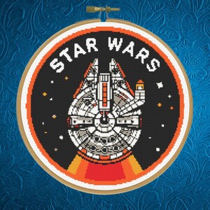Könnte beinhalten: Ein runder Stickrahmen mit einem Kreuzstichmotiv des Millennium Falcon Raumschiffs aus Star Wars. Das Design zeigt einen schwarzen Hintergrund mit weißen und grauen Schiffsdetails, orangefarbenen Akzenten und dem Text STAR WARS in Weiß.
