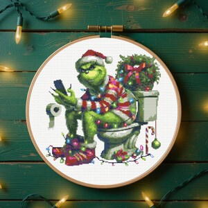 Könnte beinhalten: Kreuzstich-Stickrahmenkunst mit dem Grinch in einem Weihnachtsmannhut, der auf einer Toilette sitzt und ein Smartphone benutzt. Der Grinch ist mit Weihnachtsbeleuchtung und einem gestreiften Hemd geschmückt. Ein Weihnachtskranz und eine Zuckerstange sind ebenfalls vorhanden.