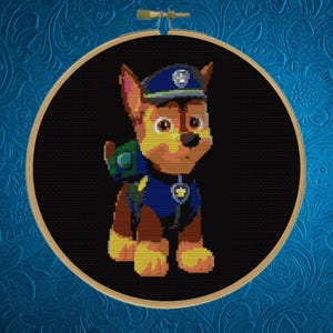 May include: A circular wooden embroidery hoop containing a cross-stitch pattern of a brown and tan German Shepherd puppy character wearing a blue police uniform, a matching blue cap with a silver badge, and a green backpack on a black background.
