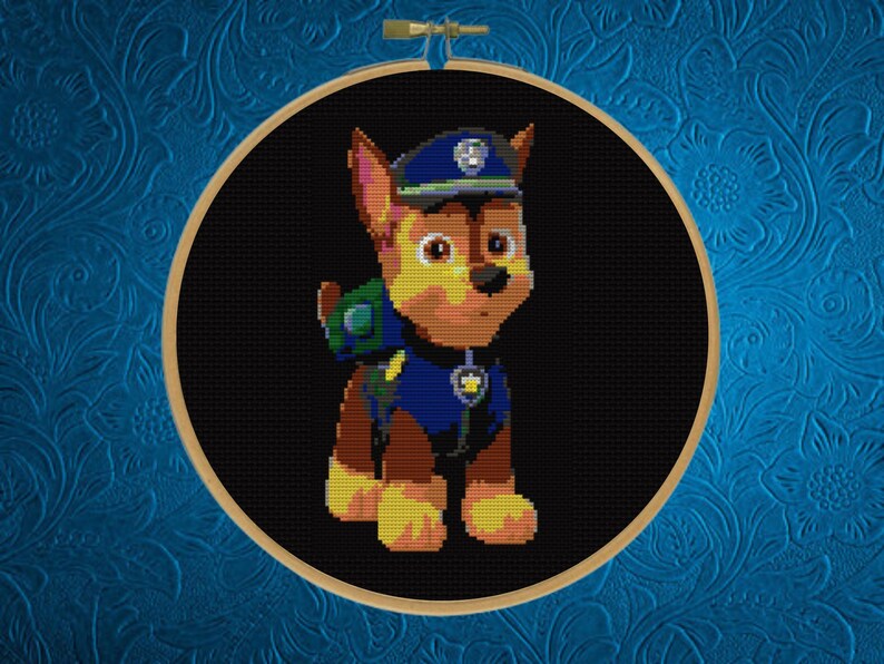 May include: A circular wooden embroidery hoop containing a cross-stitch pattern of a brown and tan German Shepherd puppy character wearing a blue police uniform, a matching blue cap with a silver badge, and a green backpack on a black background.