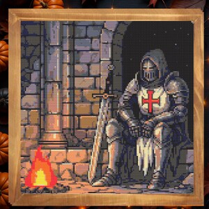 Puede incluir: Obra de arte de punto de cruz en un marco de madera que representa a un caballero con armadura completa, sentado con una espada. El caballero tiene una cruz roja en el pecho. Un peque&ntilde;o fuego arde cerca. El fondo es una pared de piedra. Calabazas y hojas de oto&ntilde;o rodean el marco.