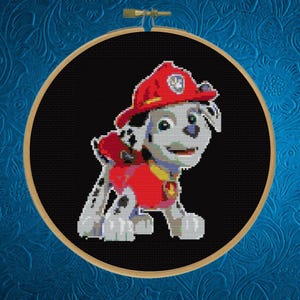 May include: A circular wooden embroidery hoop containing a cross-stitch pattern of a white and black spotted puppy character wearing a red firefighter helmet and a red vest. The character is centered against a solid black fabric background.