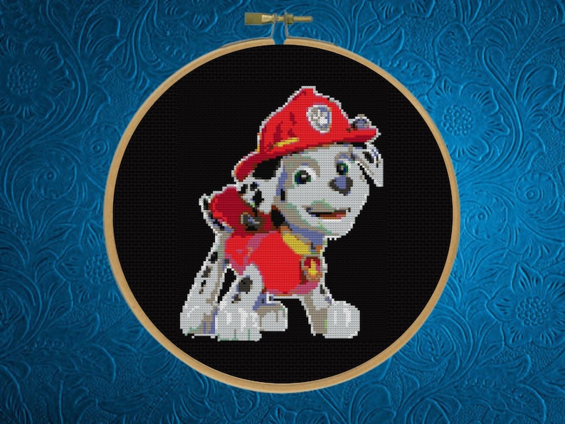 May include: A circular wooden embroidery hoop containing a cross-stitch pattern of a white and black spotted puppy character wearing a red firefighter helmet and a red vest. The character is centered against a solid black fabric background.