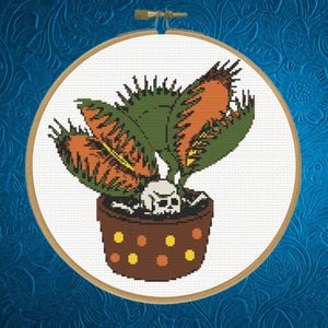 May include: Cross-stitch of a Venus flytrap in a brown pot with a skull. The plant has green leaves and orange-tipped traps. The pot has yellow and orange dots. The cross-stitch is set in a wooden hoop against a blue background.