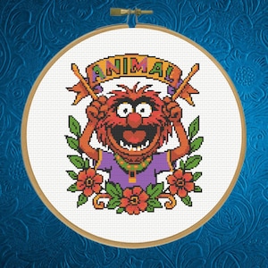 May include: A cross-stitch hoop featuring the Muppet character Animal. The design includes Animal's face, the word "ANIMAL" in a banner, and floral accents. The color palette includes red, orange, purple, and green.
