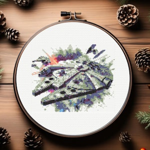 May include: A circular wooden embroidery hoop containing a cross-stitch pattern of a grey spaceship resembling the Millennium Falcon. The design features pixelated details in shades of grey, blue, and purple, with a small black fighter craft above.