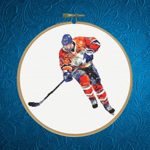 Op de afbeelding: Een kruissteek borduurwerk in een ronde houten borduurring, met een ijshockeyspeler in een oranje, blauw en wit uniform die vooruit schaatst met een zwarte hockeystick op een witte stoffen achtergrond.