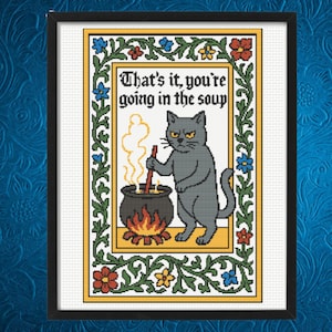 Può includere: Opera d'arte a punto croce incorniciata in nero, raffigurante un gatto grigio che mescola un calderone sopra un fuoco. Il testo recita "That's it, you're going in the soup" in un font in stile medievale, circondato da un bordo floreale.