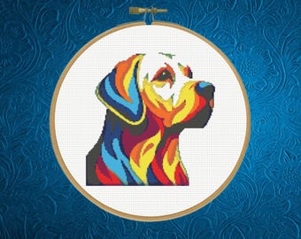 May include: A cross-stitch pattern of a dog profile in a circular wooden embroidery hoop. The dog is rendered in a vibrant, multi-colored palette featuring bold blocks of blue, yellow, red, and black against a plain white fabric background.