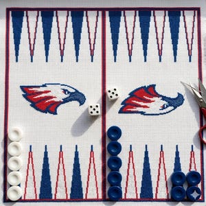 May include: A white needlepoint canvas featuring a backgammon board layout with blue and red triangular points. Two eagle head motifs are stitched in the center. The canvas includes white and blue game checkers, two dice, and metal scissors.