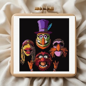 May include: A square cross-stitch pattern featuring four Muppet characters against a black background, framed in a light wood embroidery hoop. The design displays Dr. Teeth, Janice, Animal, and Floyd Pepper in a colorful, pixelated art style.