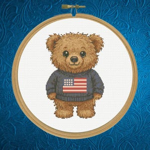 May include: A cross-stitch design of a brown teddy bear wearing a navy blue sweater with the American flag on the front. The bear is centered in a wooden embroidery hoop against a white background.