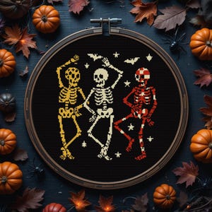 May include: A cross-stitch design of three dancing skeletons in a wooden embroidery hoop. The skeletons are in yellow, white, and red, with bats and stars. The background is black, surrounded by pumpkins and fall leaves.