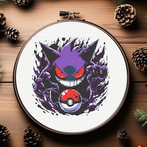 May include: A circular wooden embroidery hoop containing a cross-stitch pattern of a purple Gengar character with red eyes and sharp teeth, holding a red and white Poké Ball, surrounded by a dark purple and black pixelated aura effect.
