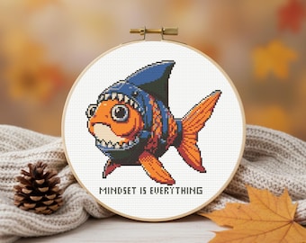 May include: A cross-stitch hoop featuring an orange fish wearing a blue shark fin hat. The fish has a toothy grin and large eyes. The text "MINDSET IS EVERYTHING" is below the fish. The hoop is set against a blurred autumn background.