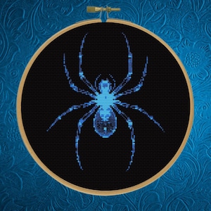 Könnte beinhalten: Ein Kreuzstich-Design einer Spinne in einem hölzernen Stickrahmen. Die Spinne ist blau auf schwarzem Hintergrund. Der Hintergrund ist ein strukturiertes, dunkelblaues Gewebe mit floralem Muster.