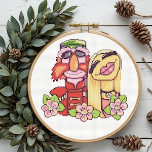 May include: A cross-stitch embroidery in a wooden hoop featuring a pixel-art style illustration of two characters. One character has a red uniform and a green hat, while the other has long blonde hair. Pink flowers are at the base of the figures.