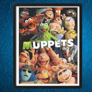 Op de afbeelding: Ingelijste kruissteek met een collage van Muppet-personages, waaronder Kermit de Kikker. Het woord "MUPPETS" is in groen en wit gestikt. Het kunstwerk is ingelijst in een zwarte lijst.
