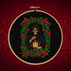 May include: Cross-stitch embroidery of the Grim Reaper holding a lantern, surrounded by a wreath of red roses and green leaves. The design is set against a black background within a wooden embroidery hoop.