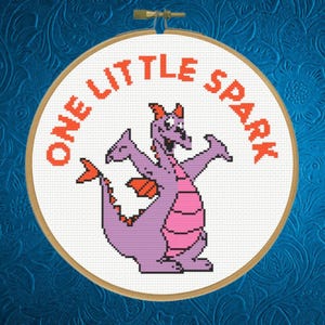 May include: Cross-stitch embroidery in a wooden hoop featuring a purple dragon with pink accents. The text "ONE LITTLE SPARK" is in orange around the top of the hoop. The background is a textured blue.