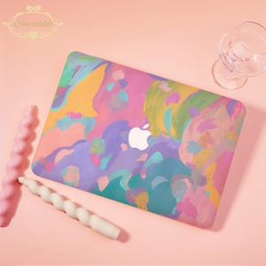 May include: A laptop case with an abstract painted design in pink, purple, yellow, and teal. The Apple logo is visible. Two spiral candles and a wine glass are on a pink surface. The case has a colourful, artistic design.