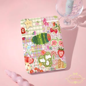 May include: A patterned tablet case with a patchwork design. The case features illustrations of a green caterpillar, strawberries, flowers, and a star. The design incorporates various patterns and colours, including pink, green, and red.