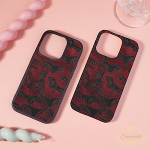 Puede incluir: Dos fundas rectangulares para teléfono con un fondo rojo oscuro y un patrón repetitivo de murciélagos dibujados con líneas negras. Cada funda tiene un gran recorte cuadrado en la parte superior para la lente de la cámara.