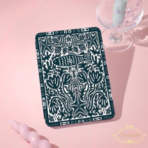 May include: A rectangular e-reader cover featuring a dark teal and white pattern of fish, starfish, and aquatic plants. The design includes a border of small arrows and floral motifs. The surface is flat with a repeating, symmetrical illustration.