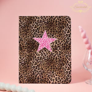 Può includere: Un quaderno con una copertina con stampa leopardata e una stella rosa con un motivo a cuore. Il quaderno è rettangolare con angoli arrotondati. La stella è centrata sulla copertina. Lo sfondo è rosa.