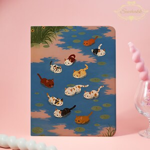 May include: A rectangular notebook featuring an illustration of ten small cats swimming in a blue pond with green lily pads. The cats are various colors including orange, brown, white, and black. The background is a soft pink color.