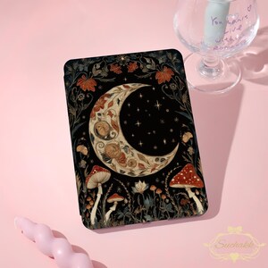 May include: A black tablet case featuring a crescent moon design with floral and mushroom accents. The moon is cream-colored with intricate details, set against a starry night sky. Red and white mushrooms are at the bottom.