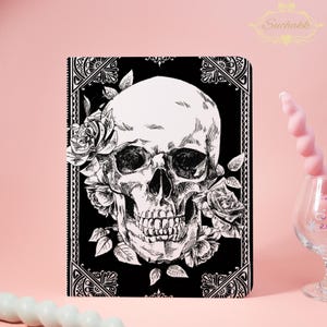 Pode incluir: Caderno preto com um desenho de caveira branca adornada com rosas. O design é emoldurado por uma borda ornamentada. O logotipo Suchakk é visível no canto superior direito. O design é gótico.