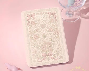 May include: A light pink phone case with a floral design featuring roses, bows, and doves. The case has a decorative border and a heart-shaped design. A pink candle and a wine glass with text are also in the image.