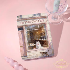 May include: A rectangular print featuring a cat cafe scene with the text "Le Petit Chat Café". The image shows a white cat with a pink bow, a tabby cat, and a cafe interior with tables and chairs. The color palette includes pink, beige, and brown.