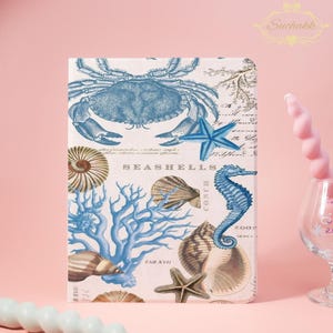 Puede incluir: Una funda decorativa para tablet con diseño náutico. Presenta un cangrejo azul, una estrella de mar, un caballito de mar y varias conchas. El diseño incluye ilustraciones botánicas y la palabra "SEASHELLS" sobre un fondo crema.
