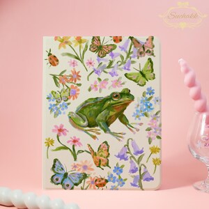 Op de afbeelding: Een crèmekleurige tablethoes met een levendige illustratie van een groene kikker, vlinders en kleurrijke bloemen. Het ontwerp bevat roze, blauwe en gele bloemelementen, met een lieveheersbeestje. Het Suchakk-logo staat rechtsboven.