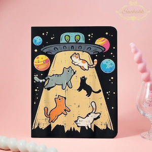 May include: A black notebook cover with a cartoon illustration of cats being beamed up by a UFO. The design features a black space background with planets and stars, and a light beam shining on the cats. The UFO has two green alien eyes.