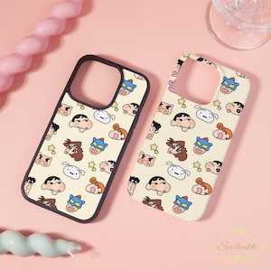 May include: Two phone cases with a repeating cartoon character pattern. The cases have a cream-colored background with various cartoon faces in different colors. One case has a black border, and the other has a cream-colored border. The cases are on a pink surface.