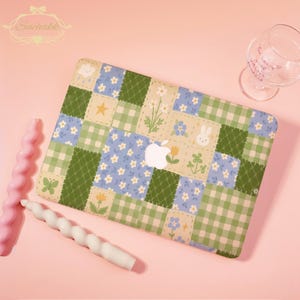 May include: A laptop case with a patchwork design featuring green, blue, and cream-colored squares. The case has floral, star, and bunny graphics. The Apple logo is visible. The background is pink, with two spiral candles and a wine glass.