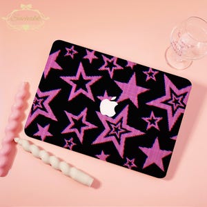 May include: Black laptop case featuring a pink star pattern. The Apple logo is in the centre. The case is on a pink surface, with two spiral candles and a wine glass nearby.