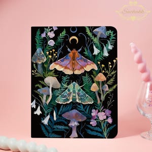 Pode incluir: Um caderno retangular com capa preta apresentando uma ilustração colorida de duas mariposas, vários cogumelos, samambaias e pequenas flores. Uma lua crescente dourada está posicionada acima da mariposa superior.