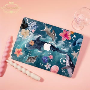 May include: A decorative laptop skin featuring a vibrant ocean scene with whales, starfish, and flowers. The design includes a whale shark with a flower on its head, set against a teal background with white wave details. The skin is surrounded by pink and white candles.