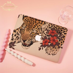 Könnte beinhalten: Eine Laptop-Hülle mit einer detaillierten Illustration eines Leoparden mit bernsteinfarbenen Augen, umgeben von dunkelroten Blumen und grünen Blättern. Das Design ist auf beigem Hintergrund und enthält das weiße Apple-Logo.