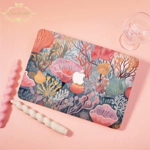 May include: A laptop case with a vibrant coral reef pattern in shades of pink, orange, and blue. The case displays a white Apple logo. It is accompanied by pink and white spiral candles and a wine glass. The Suchakk logo is visible in the top left corner.