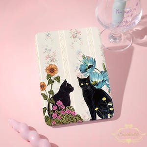 May include: A rectangular e-reader case featuring a cream-colored background with vertical stripes and floral patterns. Two black cats are depicted, one sitting among pink flowers and a sunflower, and the other near large blue flowers.