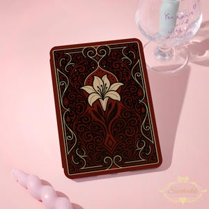 May include: A rectangular e-reader case featuring a dark red background with intricate black and cream-colored swirling filigree patterns and a central cream-colored lily flower illustration. The case has a smooth, flat surface design.