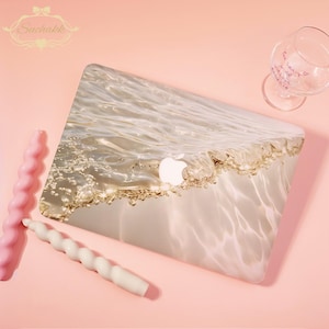 May include: A laptop case with a water-like design, featuring the Apple logo. The case has a gold and white wave pattern. Two spiral candles, one pink and one white, and a wine glass are on a pink surface.