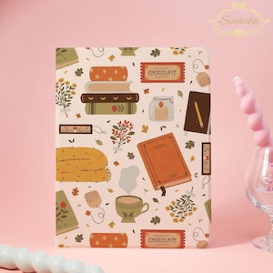May include: A decorative notebook with a cream background featuring illustrations of books, a cup of tea, a scarf, and the word "CHOCOLATE" on a rectangular label. The design includes autumn-themed elements in shades of orange, green, and brown.