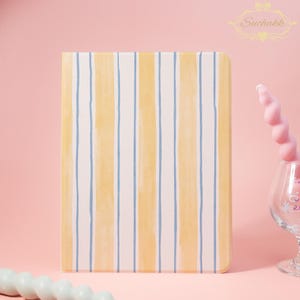 May include: A rectangular object with a striped pattern in yellow, white, and blue. The stripes run vertically. The object is set against a pink backdrop. A pink spiral candle and a glass are visible. The Suchakk logo is in the top right corner.