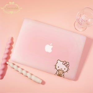 May include: Pink laptop with a white Apple logo and a Hello Kitty sticker holding a teddy bear. Two spiral candles, one pink and one white, and a wine glass are also on the pink surface.
